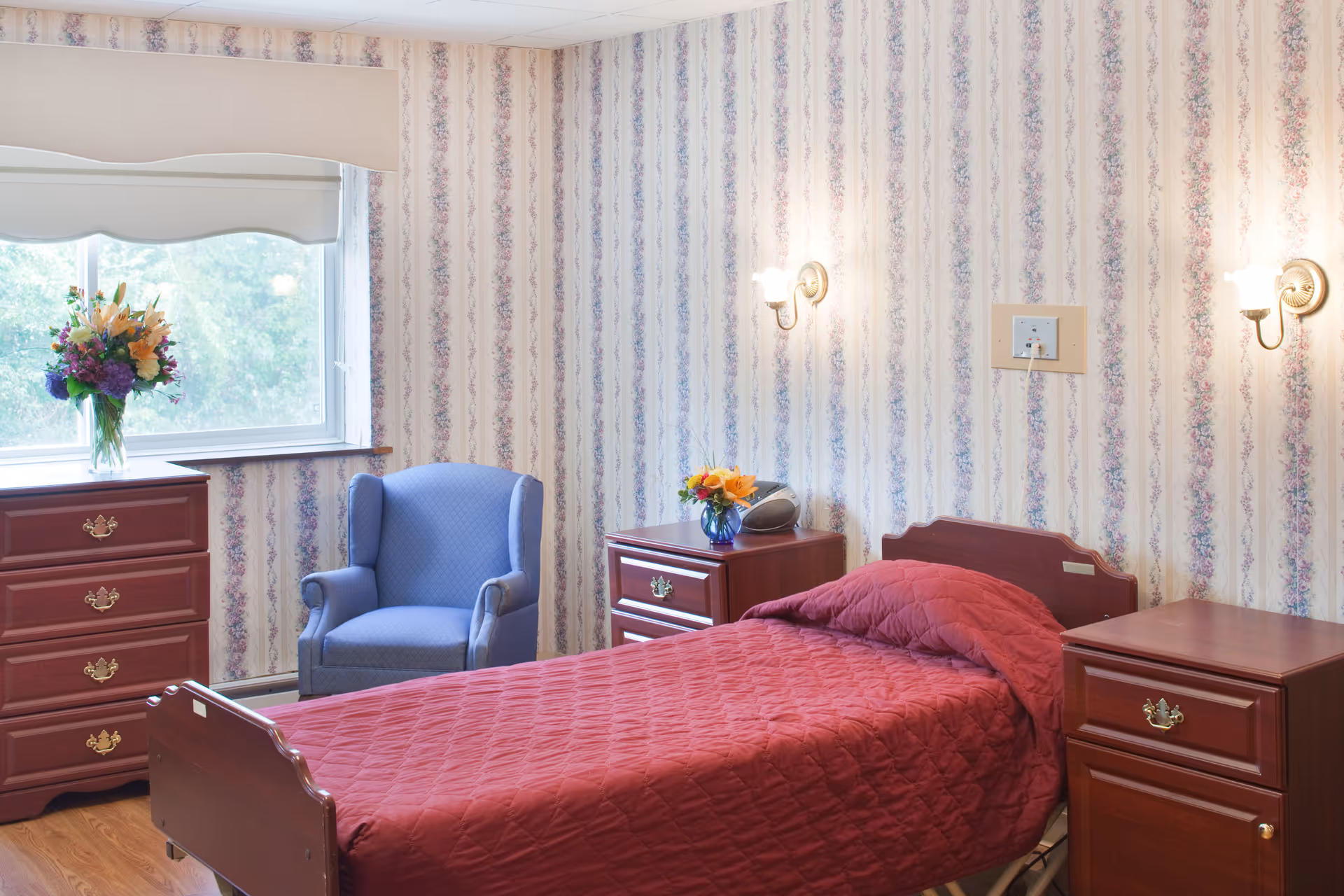 A bedroom in a senior living facility with a single bed covered in a red quilted blanket, two wooden nightstands with brass handles, a blue upholstered armchair, a wooden dresser, floral wallpaper, and a window with a floral bouquet on the windowsill.