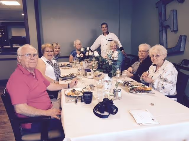 A group of seven elderly people and one standing staff member gathered around a dining table with plates of food, drinks, and a flower centerpiece in a dining room setting.