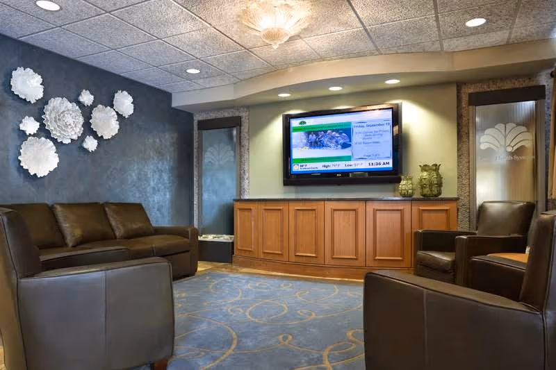 Comfortable seating area with leather sofas and chairs facing a wall-mounted TV above wooden cabinetry, decorative wall art, and patterned carpet.