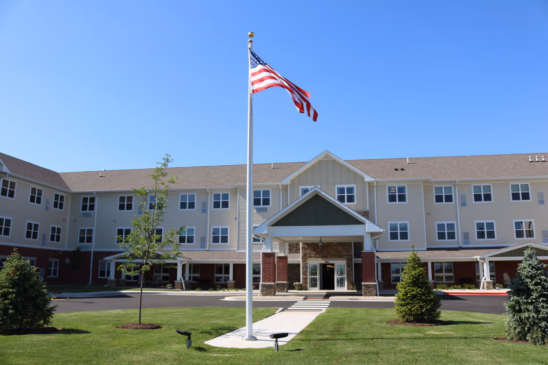 Front exterior view of Heritage Woods of Minooka, a three-story building with beige siding and brick accents. A flagpole with an American flag is centered in front of the entrance, surrounded by a well-maintained lawn and small trees under a clear blue sky.
