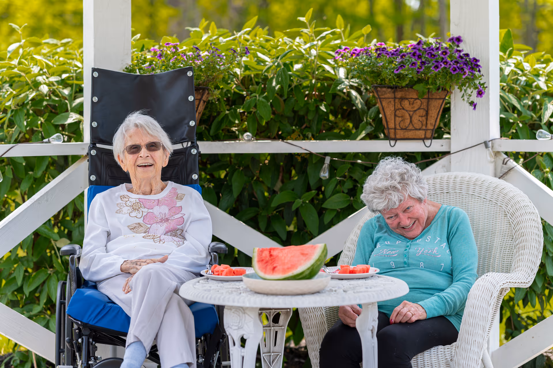 Two elderly women sitting outdoors on a porch or patio. One woman is in a wheelchair wearing sunglasses and a white sweater with floral designs, smiling. The other woman is sitting in a white wicker chair wearing a teal long-sleeve shirt, also smiling and looking down. A white round table between them holds a large slice of watermelon and plates with watermelon pieces. Green foliage and purple flowers in hanging baskets are visible in the background.