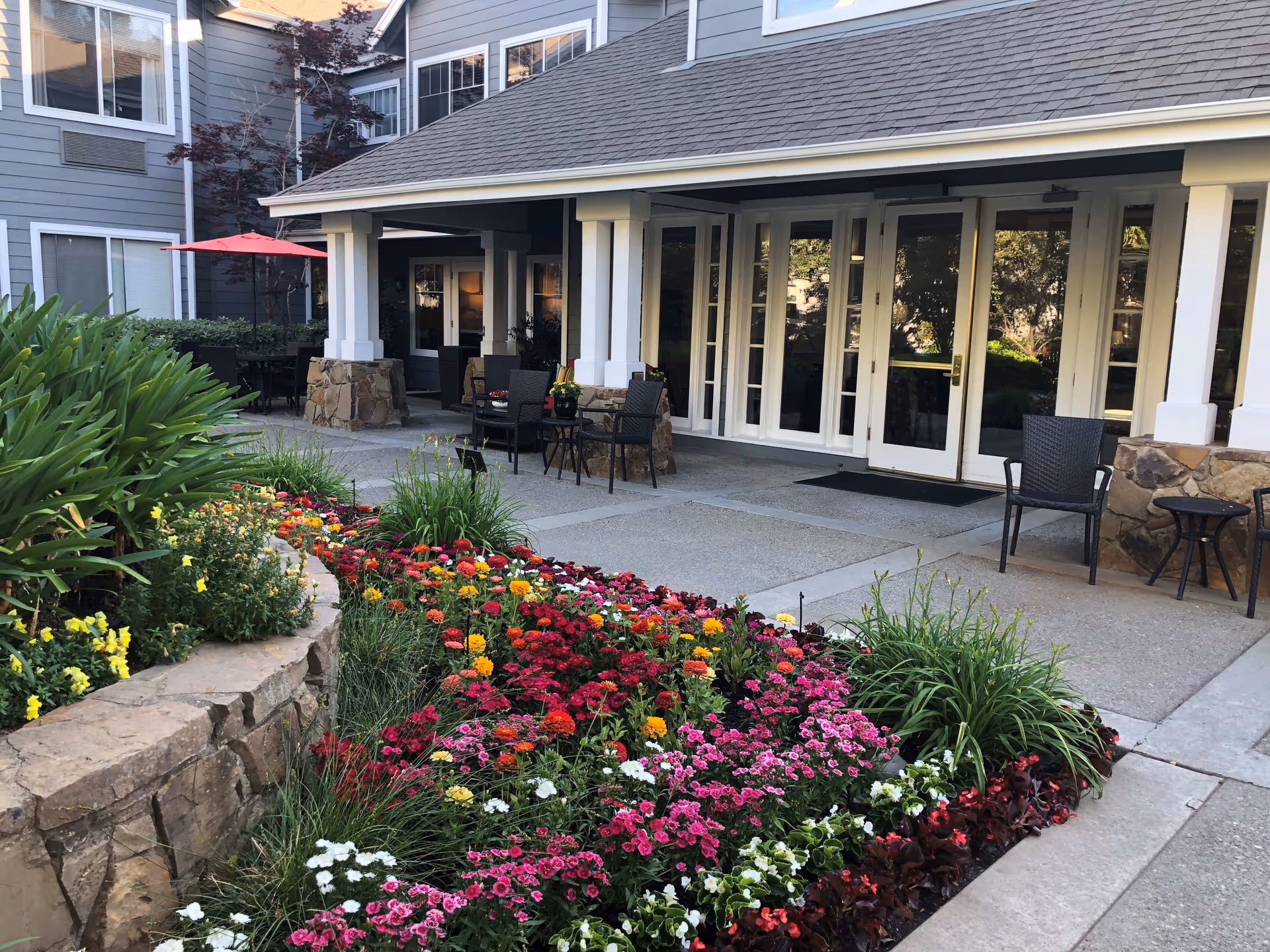 Outdoor patio area of a senior living facility with a colorful flower bed in the foreground, stone-bordered garden, several black chairs and small tables on a concrete patio, and a building with large windows and a covered entrance in the background.