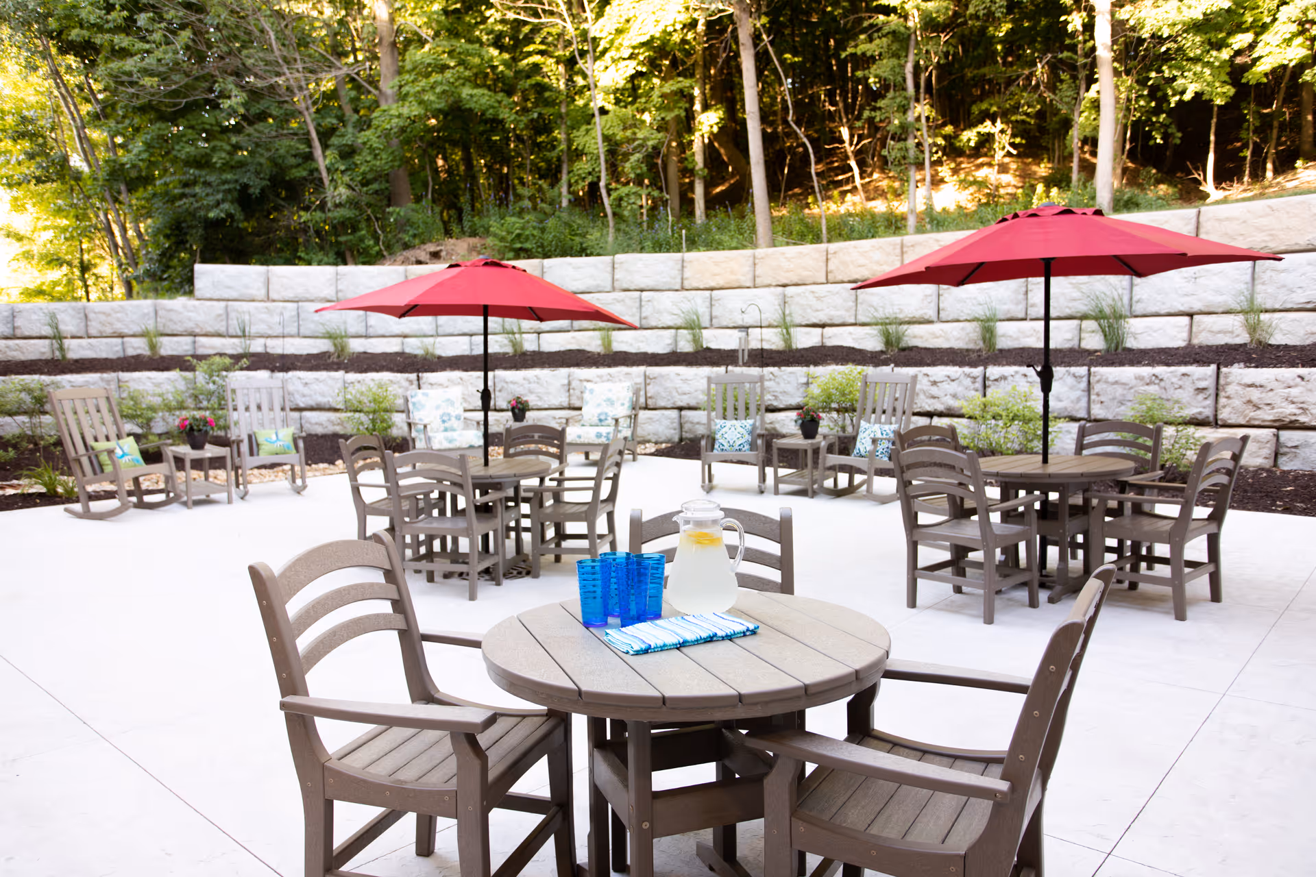 Outdoor patio area with several round wooden tables and chairs. Two tables have red umbrellas providing shade. One table in the foreground has a pitcher of lemonade and four blue glasses on a striped cloth. In the background, there are rocking chairs with cushions and small side tables, all set against a stone retaining wall with greenery and trees behind it.