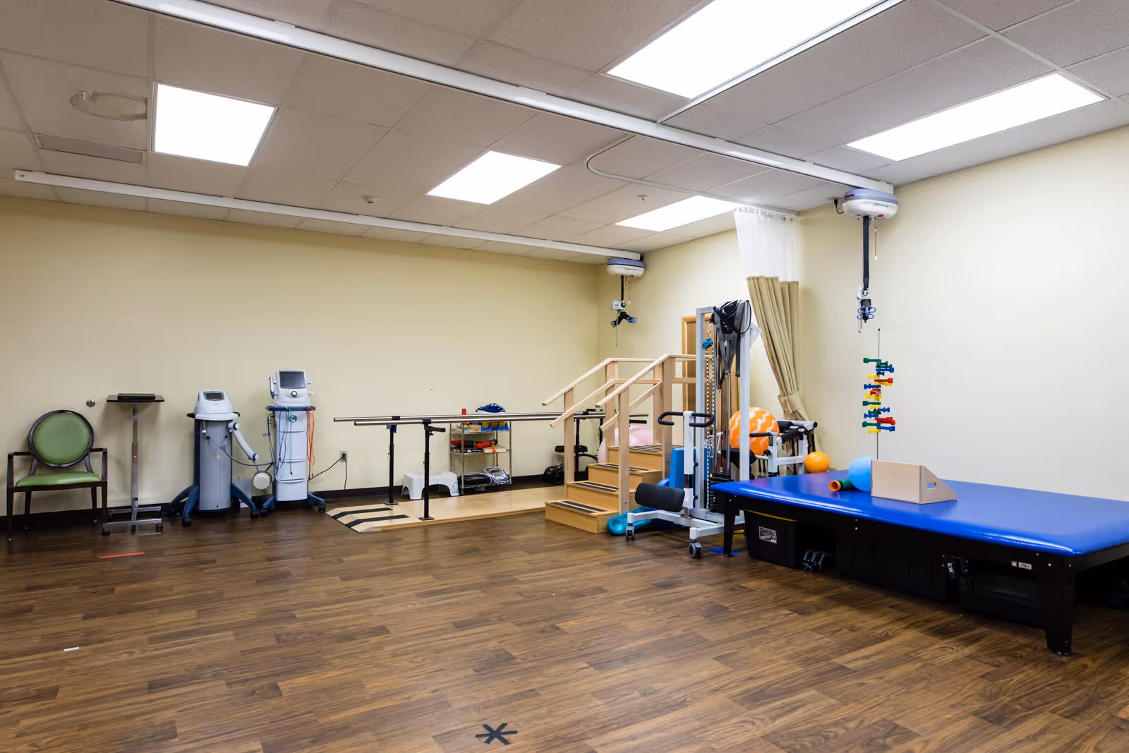 A physical therapy room with wooden flooring, parallel bars for walking exercises, a small wooden staircase, exercise equipment including therapy balls, and a padded blue therapy table. The room has beige walls and ceiling lights.