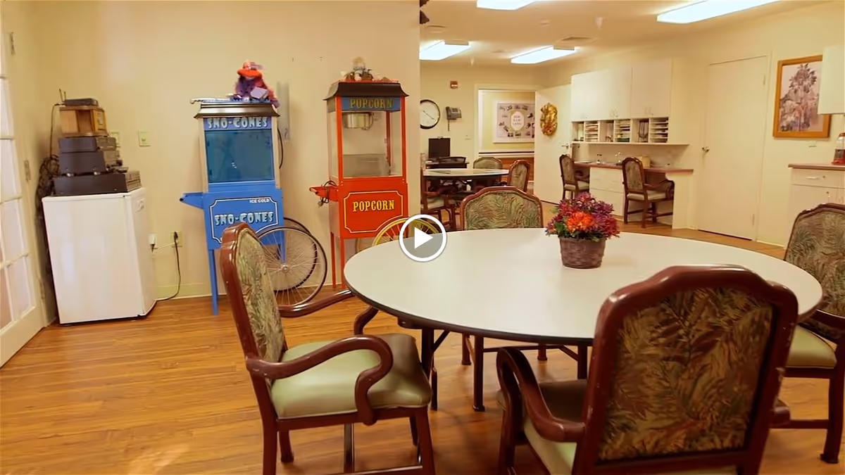 A communal room with a round table surrounded by six upholstered chairs with floral patterns. On the table is a small basket with colorful artificial flowers. In the background, there are two vintage-style machines: a blue Sno-Cones machine and a red Popcorn machine. The room has wooden flooring, cream-colored walls, and built-in desks with chairs along the far wall. A clock and framed artwork are visible on the walls.