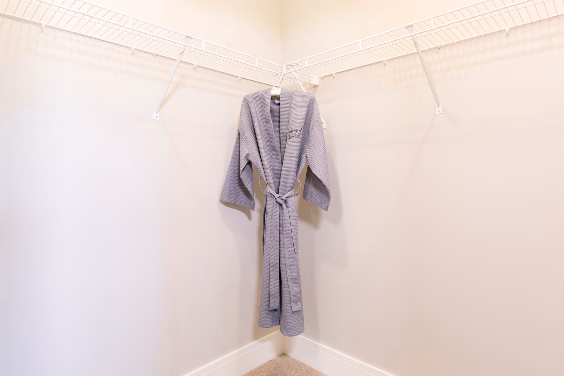 A light gray robe hanging on a white wire shelf in an empty corner closet with beige walls and carpeted floor.