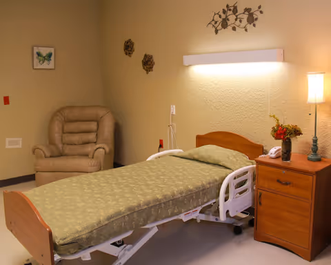 A senior bedroom with an adjustable hospital bed, wooden nightstand with a lamp and flowers, and a beige armchair against a textured wall.