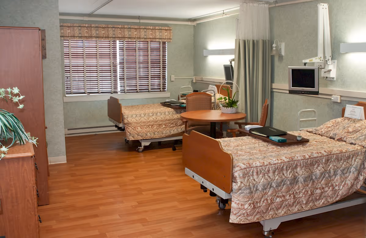 A shared senior facility bedroom with two hospital-style beds, a round table and chairs, and wood flooring.