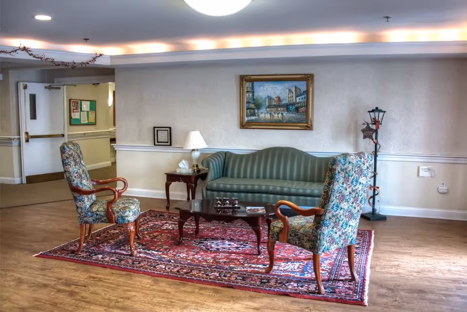 A cozy sitting area in an assisted living facility featuring a green striped sofa, two floral upholstered armchairs, a wooden coffee table on a red patterned rug, a side table with a lamp and tissues, a framed painting on the wall, and a decorative floor lamp in the corner.