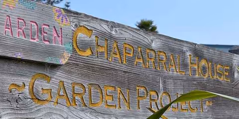 Rugged wooden sign reading Chaparral House Garden Project against a clear blue sky.