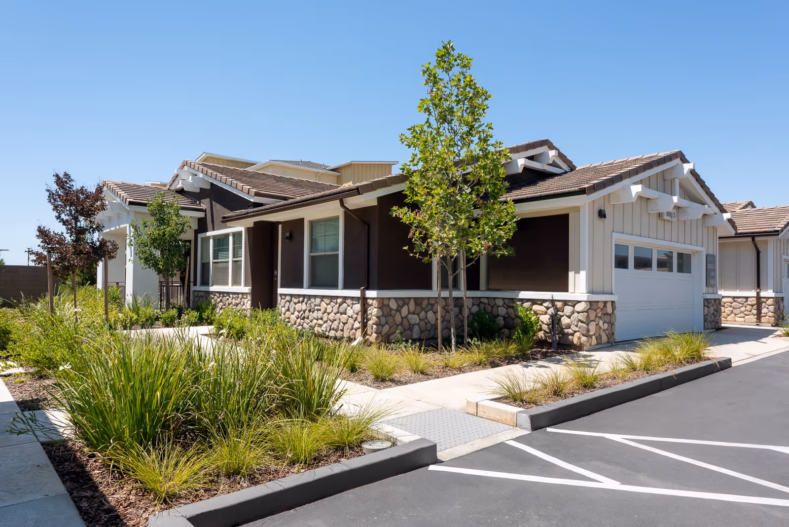 Exterior view of a single-story residential building with a stone and wood facade, surrounded by landscaped greenery and a paved parking area under a clear blue sky.