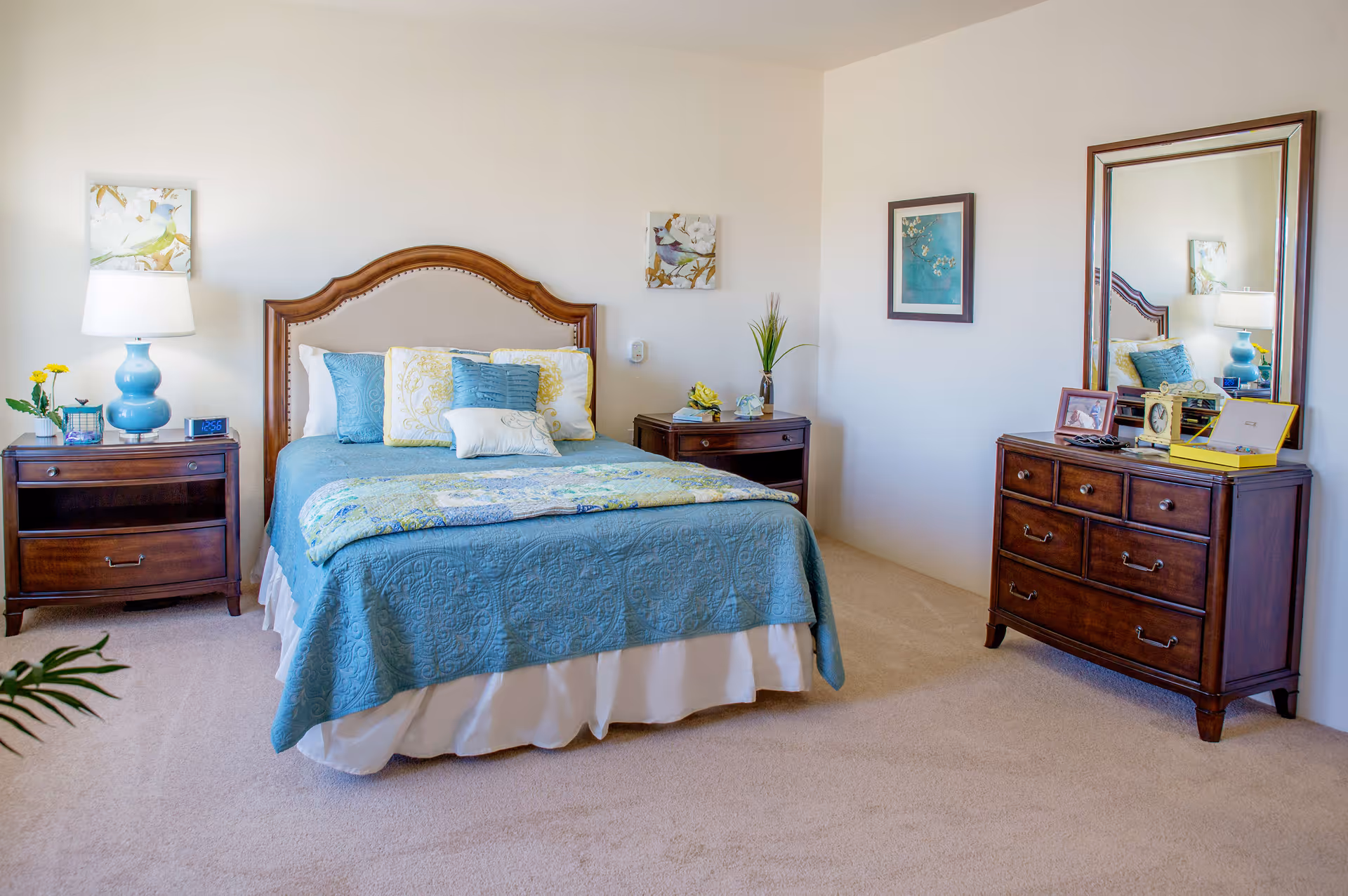 A neatly arranged bedroom with a bed featuring a wooden headboard and blue bedding with multiple pillows. There are two wooden nightstands on either side of the bed, one with a blue lamp and clock, and the other with a plant and decorative items. A wooden dresser with a large mirror and various framed photos and decor is positioned against the wall. The room has light-colored walls and carpeted flooring.