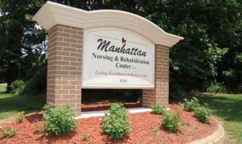 Outdoor brick sign reads Manhattan Nursing and Rehabilitation Center with mulch and shrubs.