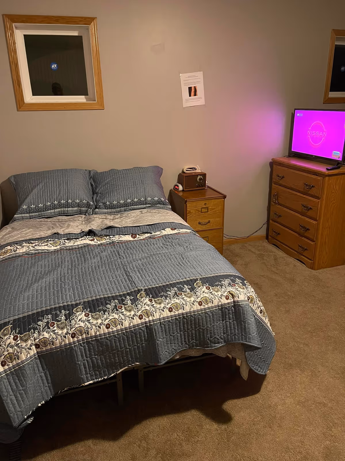 A bedroom with a bed covered in a blue and white patterned quilt and two matching pillows. There is a wooden nightstand with a vintage radio and a small clock on it next to the bed. A wooden dresser with a television on top displaying a pink screen with the Nissan logo is on the right side. The walls are plain and beige with a small window and a piece of paper taped to the wall above the nightstand.