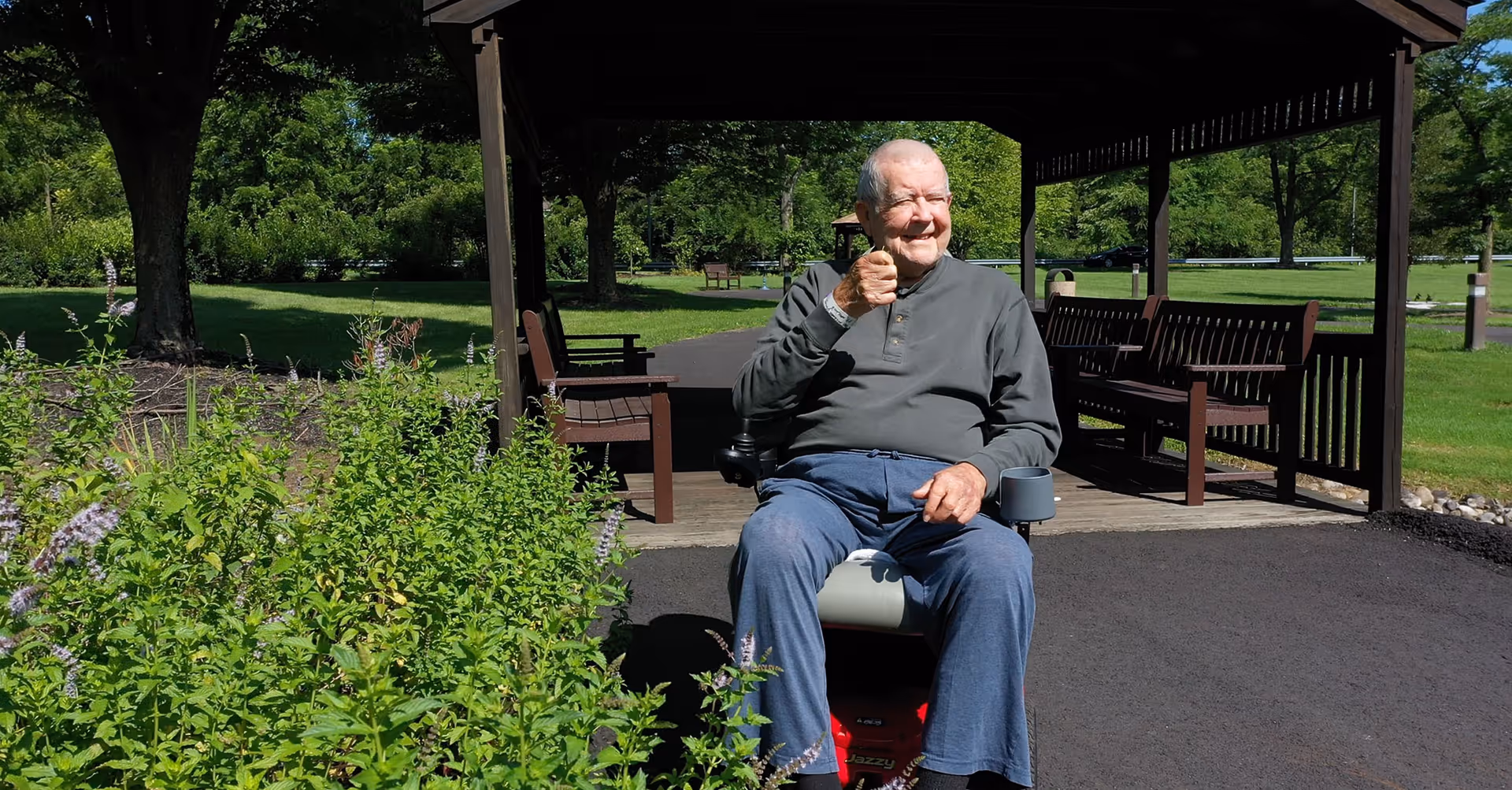 An elderly man sitting on a motorized scooter giving a thumbs-up gesture while outdoors near a wooden gazebo with benches. The area is surrounded by green grass, trees, and flowering plants under a sunny sky.