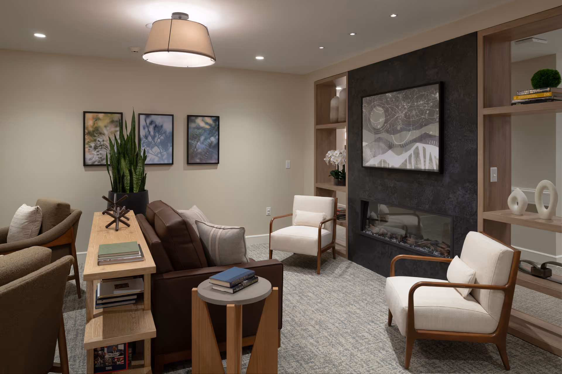 A cozy living room with a brown leather sofa, two beige armchairs with wooden arms, a wooden side table with books, and a modern black fireplace set into a dark textured wall. The room also features built-in wooden shelves with decorative items, three framed abstract art pieces on the wall, a large potted plant, and a ceiling light fixture.
