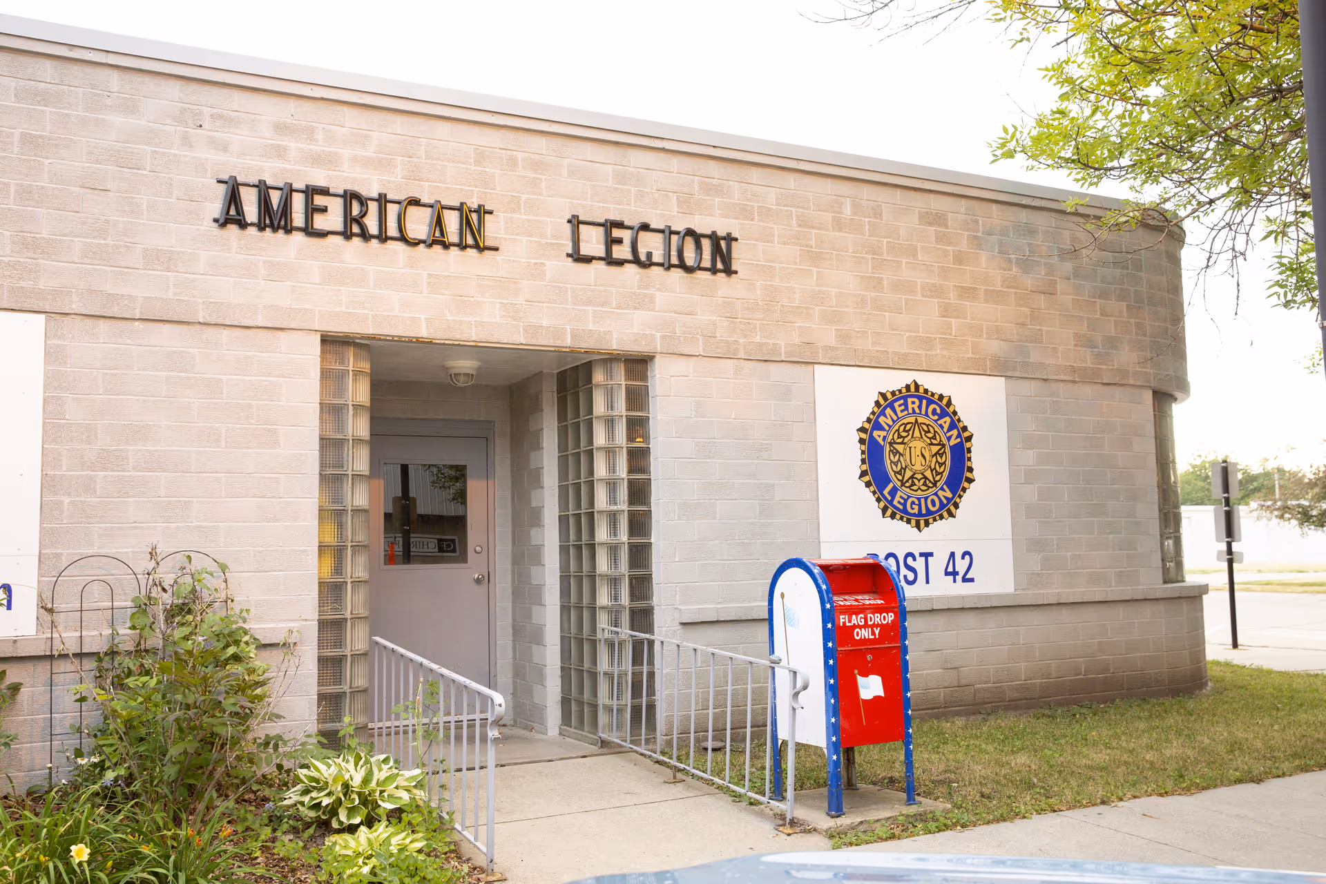 Exterior view of a building with a sign that reads 'AMERICAN LEGION' above the entrance. The entrance has a gray door flanked by glass block windows. To the right of the door is a large sign with the American Legion emblem and the text 'POST 42'. In front of the building is a red, white, and blue mailbox labeled 'FLAG DROP ONLY'. There is some greenery and a tree partially visible on the right side.
