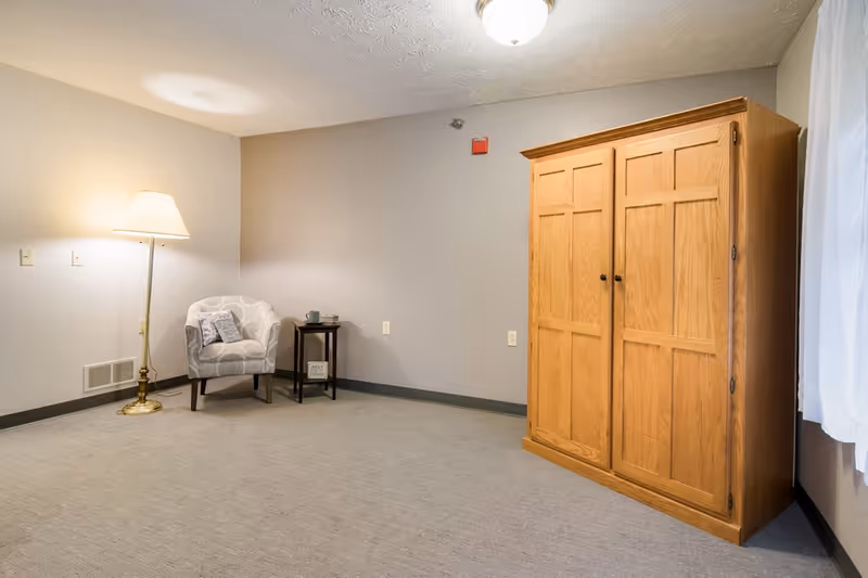 A simple room with light gray walls and carpeted floor featuring a wooden wardrobe on the right, a small round side table with a cup and a decorative item, a patterned armchair with a pillow, and a tall floor lamp providing warm light in the corner.