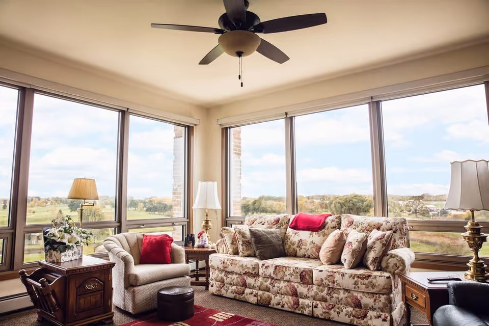 A cozy living room with large windows offering a scenic view of a green landscape. The room features a floral patterned sofa with multiple cushions, a beige armchair with a red pillow, two wooden side tables with lamps, a ceiling fan, and a small ottoman.