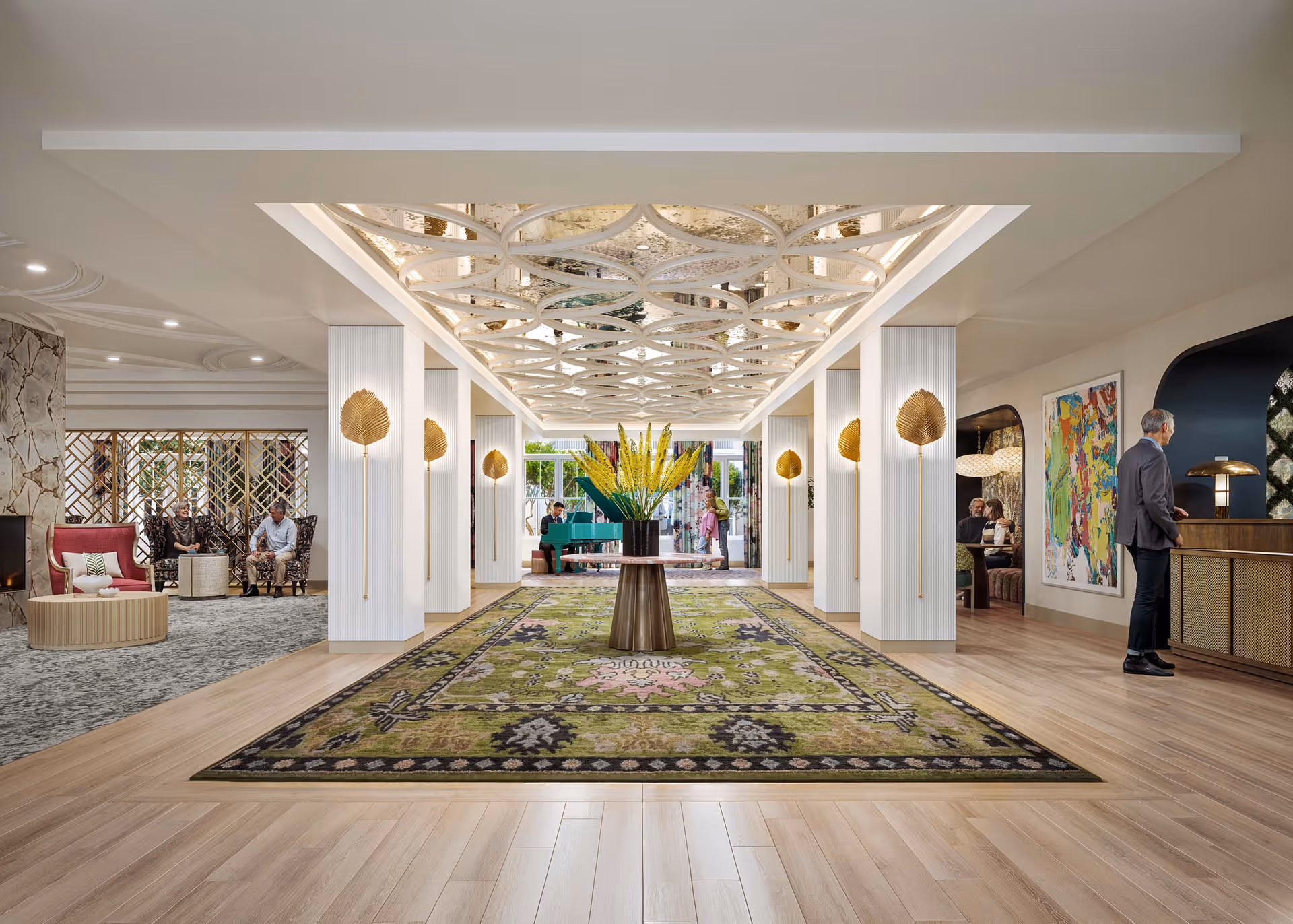 A spacious and elegant senior living facility lobby with a large green patterned rug centered under a decorative ceiling with circular designs. A round table with a tall floral arrangement is in the middle of the rug. To the left, there are seating areas with people conversing near a stone fireplace. To the right, a man stands at a reception desk with colorful artwork on the wall behind him. The far end of the lobby has large windows and a teal piano with people nearby.