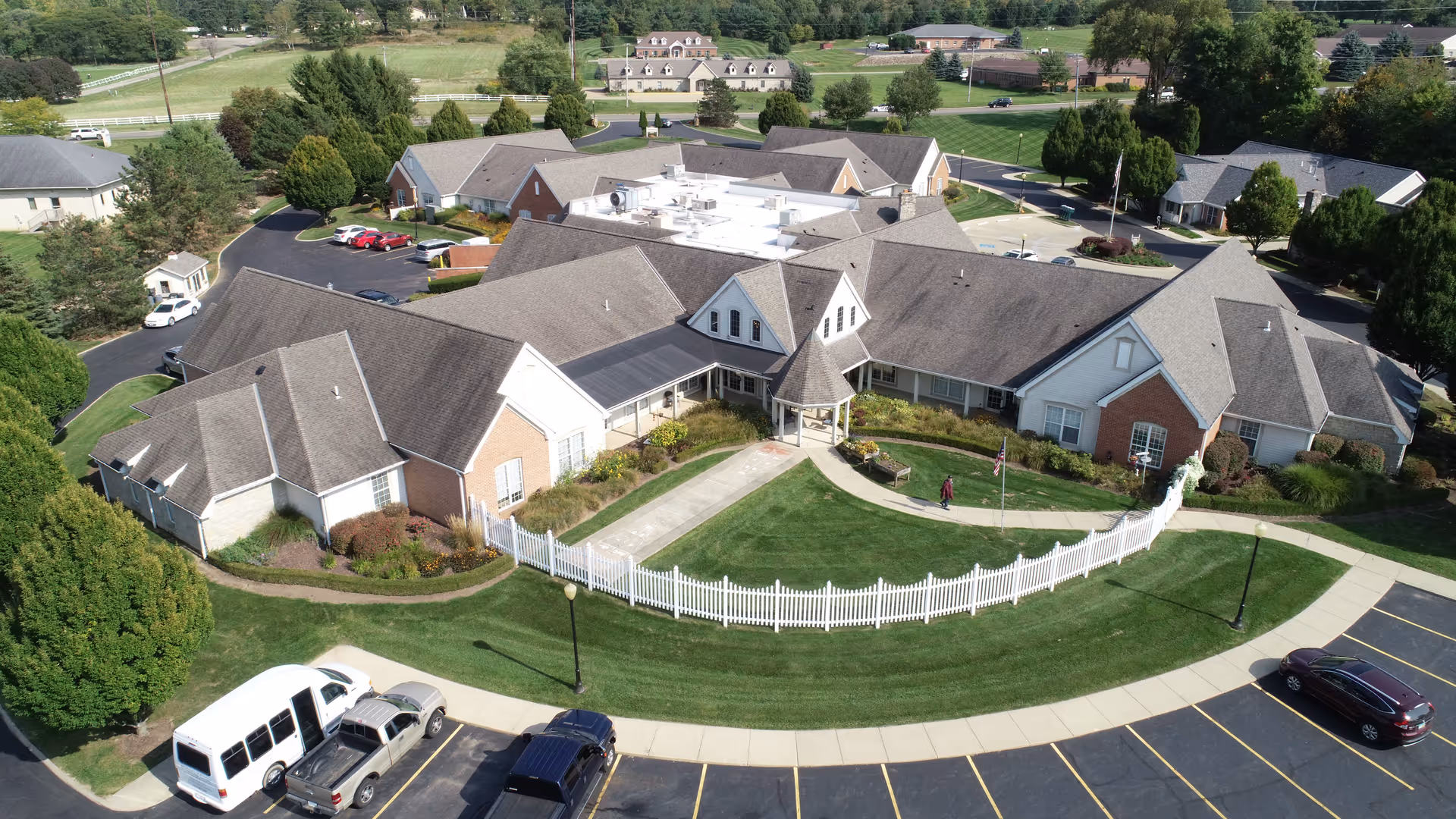 Aerial view of Wedgewood Estates senior living facility showing a large, single-story building with multiple roof sections, surrounded by well-maintained lawns, trees, and a white picket fence. Several cars are parked in the parking lot in front of the building, and a person is walking near the entrance.