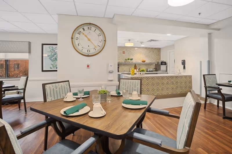 A dining area in a senior living facility with a wooden table set for four people with plates, cups, glasses, and green napkins. The room has wooden flooring, cushioned chairs with patterned upholstery, a large wall clock, and a kitchen area in the background with a patterned counter and various kitchen appliances.