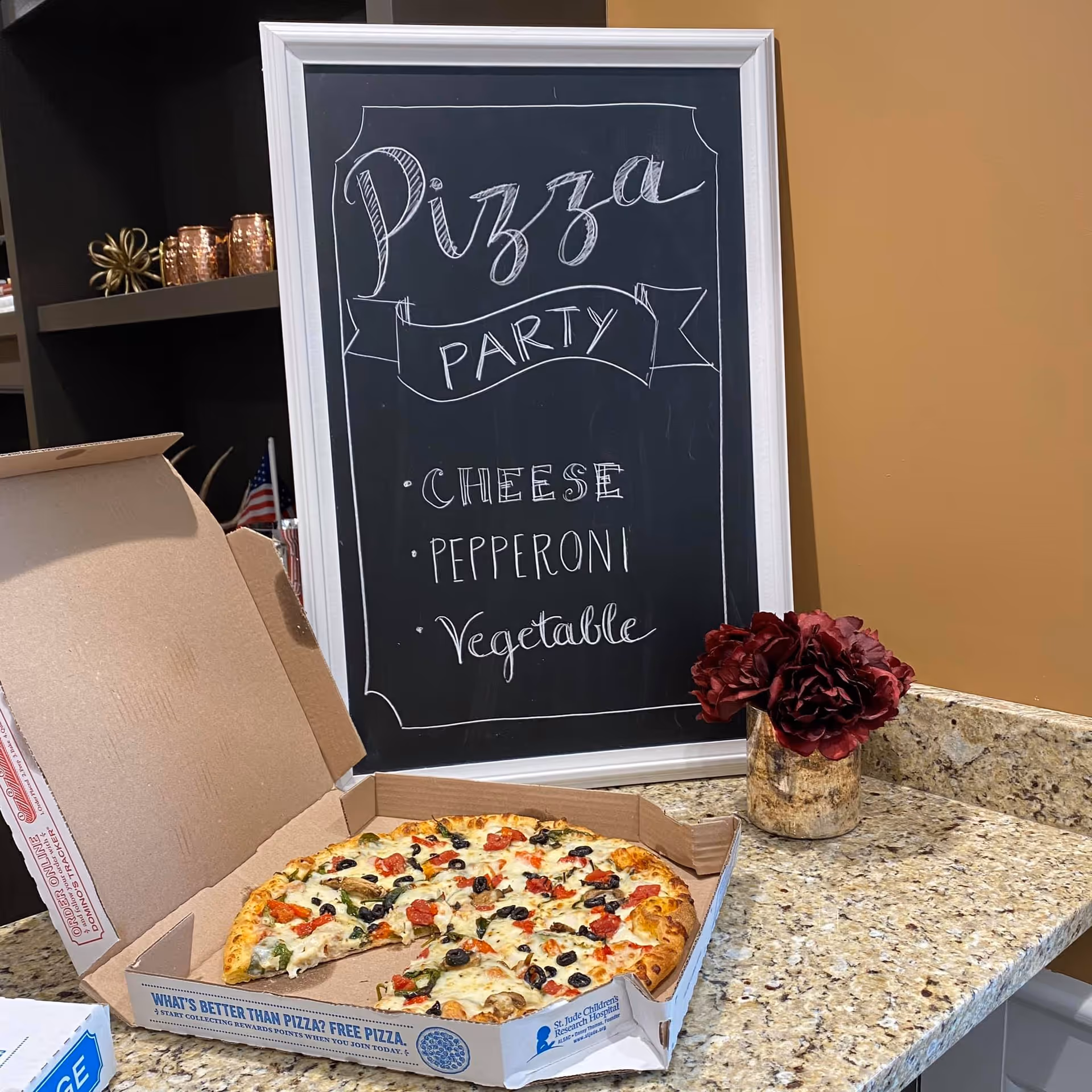 An open pizza box with a partially eaten vegetable pizza on a granite countertop. Behind it is a blackboard with white chalk writing that says 'Pizza Party' and lists cheese, pepperoni, and vegetable as options. A small vase with dark red flowers is also on the countertop.