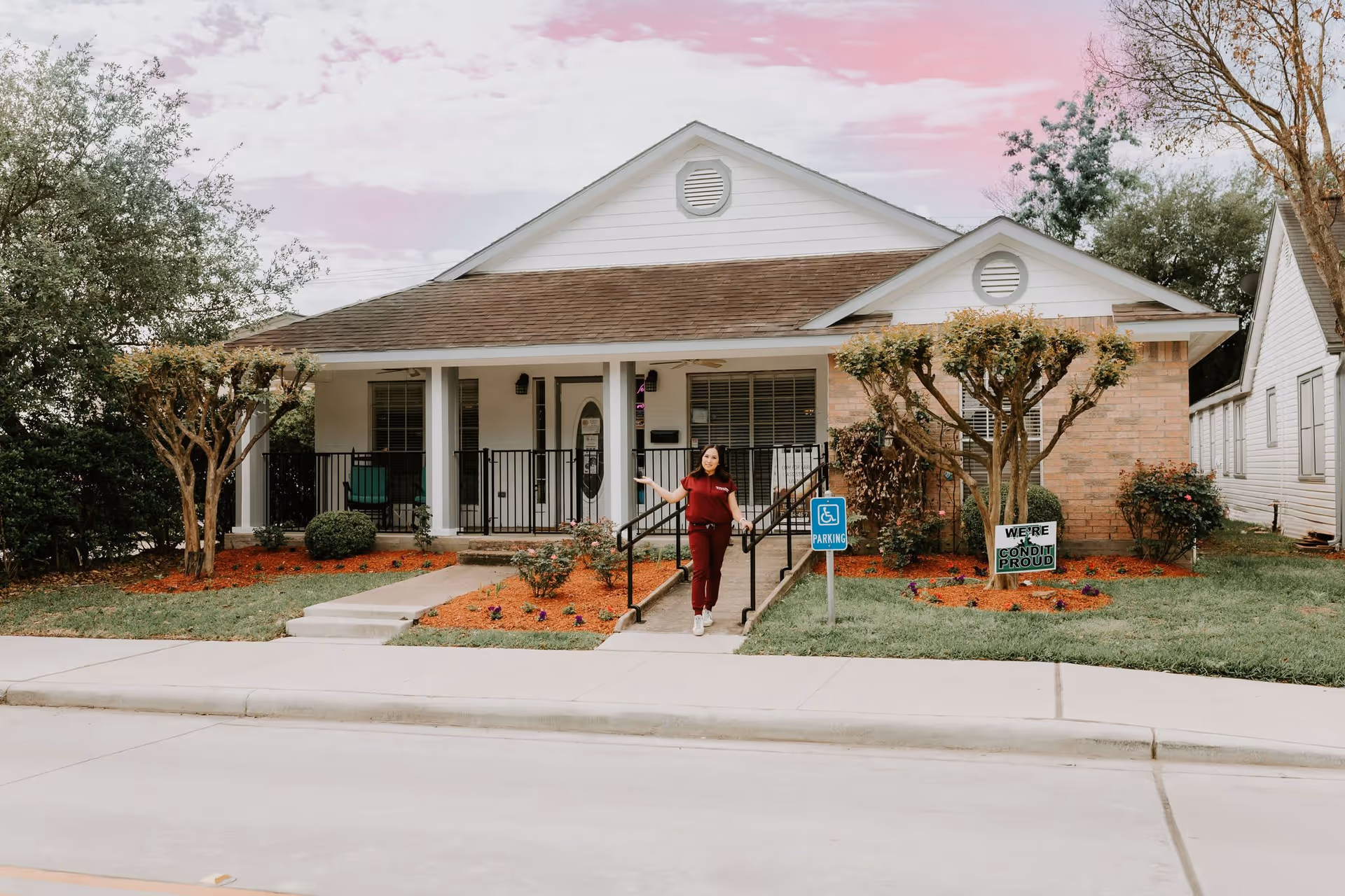 A single-story assisted living facility building with a front porch, landscaped garden, and a woman standing on the ramp leading to the entrance. The sky has a pinkish hue, and there is a handicapped parking sign and a sign that reads 'WE'RE CONDHI PROUD' in the garden area.