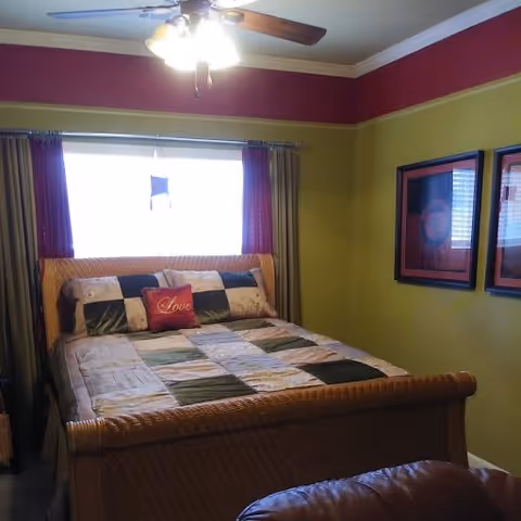 A furnished bedroom with a wooden sleigh bed, patterned bedspread, ceiling fan, and framed artwork on green-painted walls.