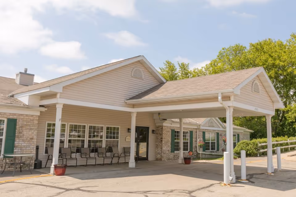 Covered main entrance of a single-story assisted living building with a porte-cochère, outdoor chairs, and a driveway.