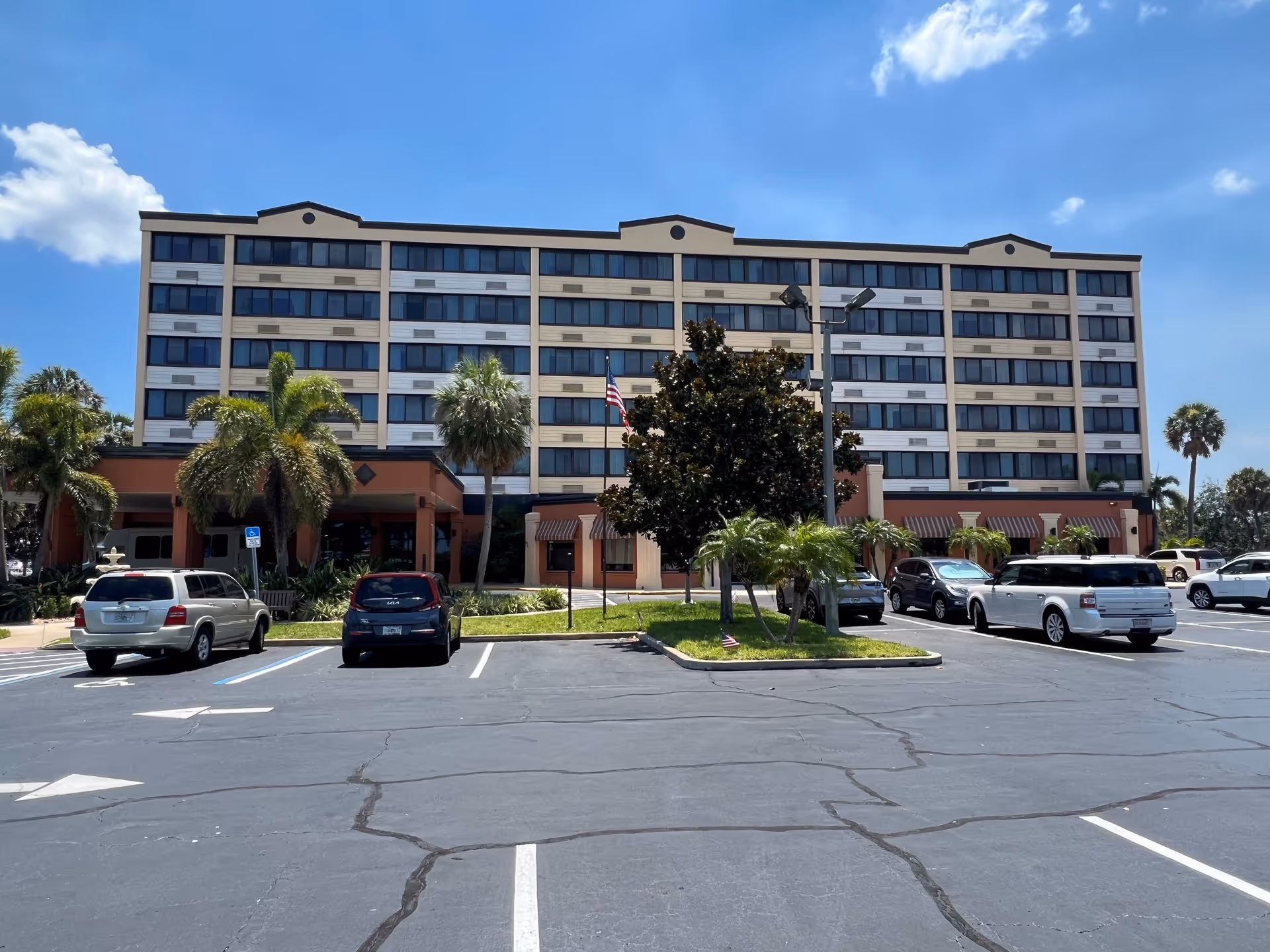 Exterior view of a multi-story senior living facility building with several windows, palm trees, and a parking lot with parked cars under a clear blue sky.