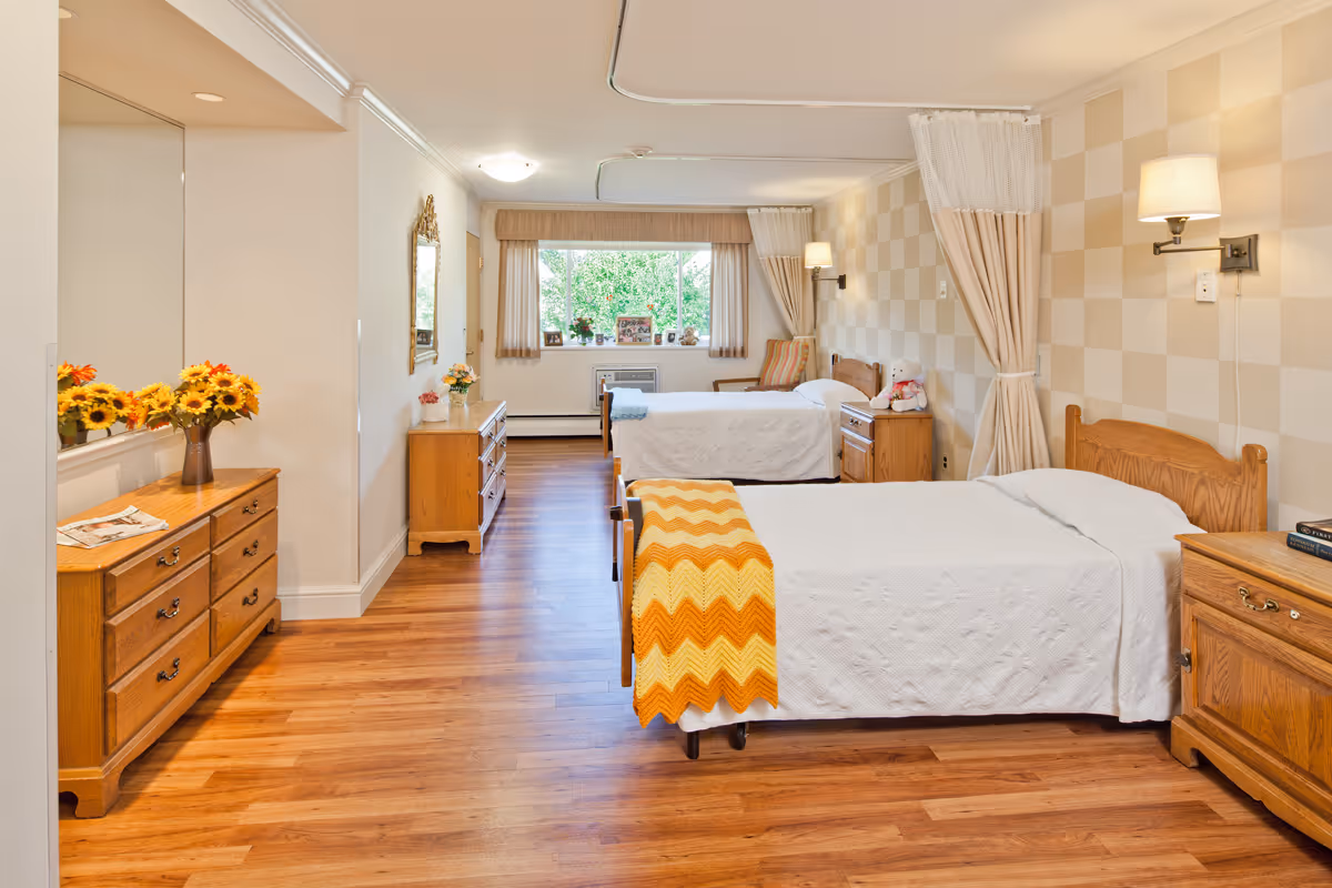 A bright and clean nursing facility bedroom with two single beds, each with white bedding and a colorful blanket at the foot. The room features wooden furniture including dressers and nightstands, a large window with curtains letting in natural light, and wall-mounted lamps. The floor is wooden, and there are decorative items such as flowers and framed photos on the furniture.