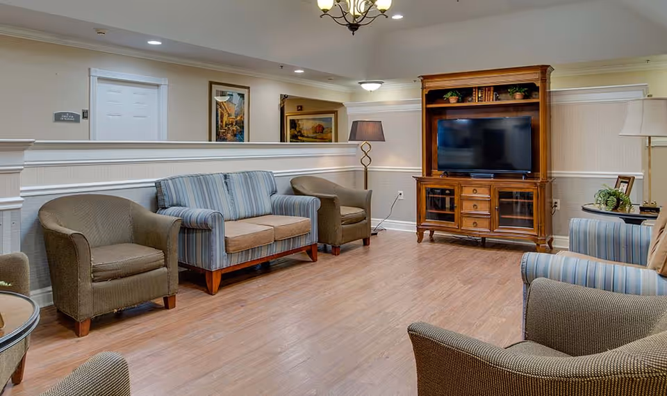 A cozy senior living facility common area with wooden flooring, a wooden TV cabinet with a flat-screen television, two striped sofas, three brown armchairs, a round side table with a lamp, and framed artwork on the walls.