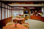 A cozy senior living common area with wooden tables and chairs, a stone fireplace with a mounted TV above it, built-in wooden shelves with decorative items, and a carpeted floor. The room has warm lighting and a comfortable, inviting atmosphere.