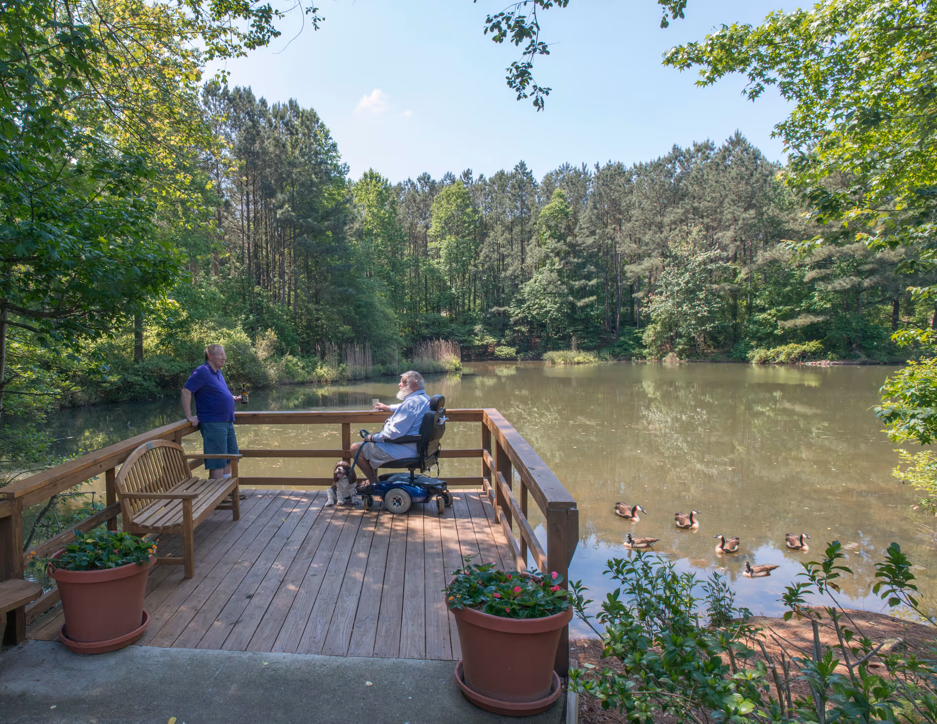 Two men, one seated in a mobility scooter, converse on a wooden deck overlooking a tree-lined pond where ducks swim nearby.
