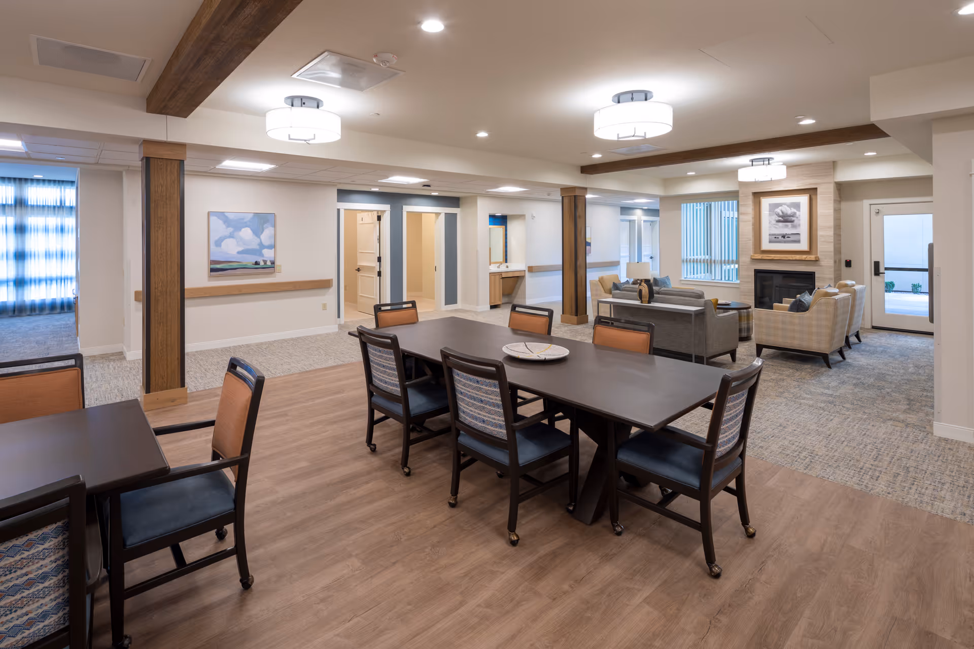 A spacious and well-lit common area in a senior living facility featuring a large dark wood dining table with eight chairs, a smaller table with two chairs, and a cozy seating area with sofas and armchairs near a fireplace. The room has wooden beams on the ceiling, modern light fixtures, and a mix of carpet and wood flooring. There are doorways and windows allowing natural light into the space.