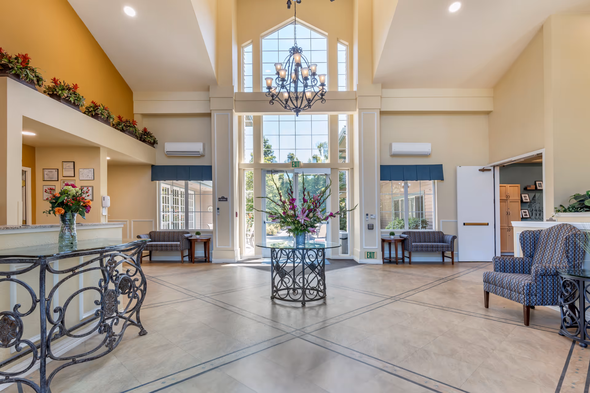 Spacious and well-lit lobby area with high ceilings and large windows allowing natural light. A decorative chandelier hangs from the ceiling. In the center, there is a round glass table with a floral arrangement. Seating areas with patterned chairs and small tables are positioned along the walls. The reception desk with a vase of flowers is visible on the left side. The overall decor is warm and inviting with neutral tones and some greenery.