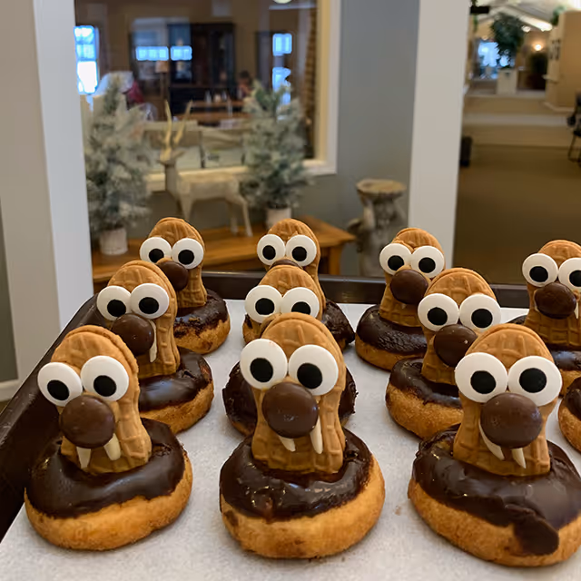 Chocolate-glazed doughnuts decorated as cookie 'walrus' faces arranged on a baking tray in a communal interior space.