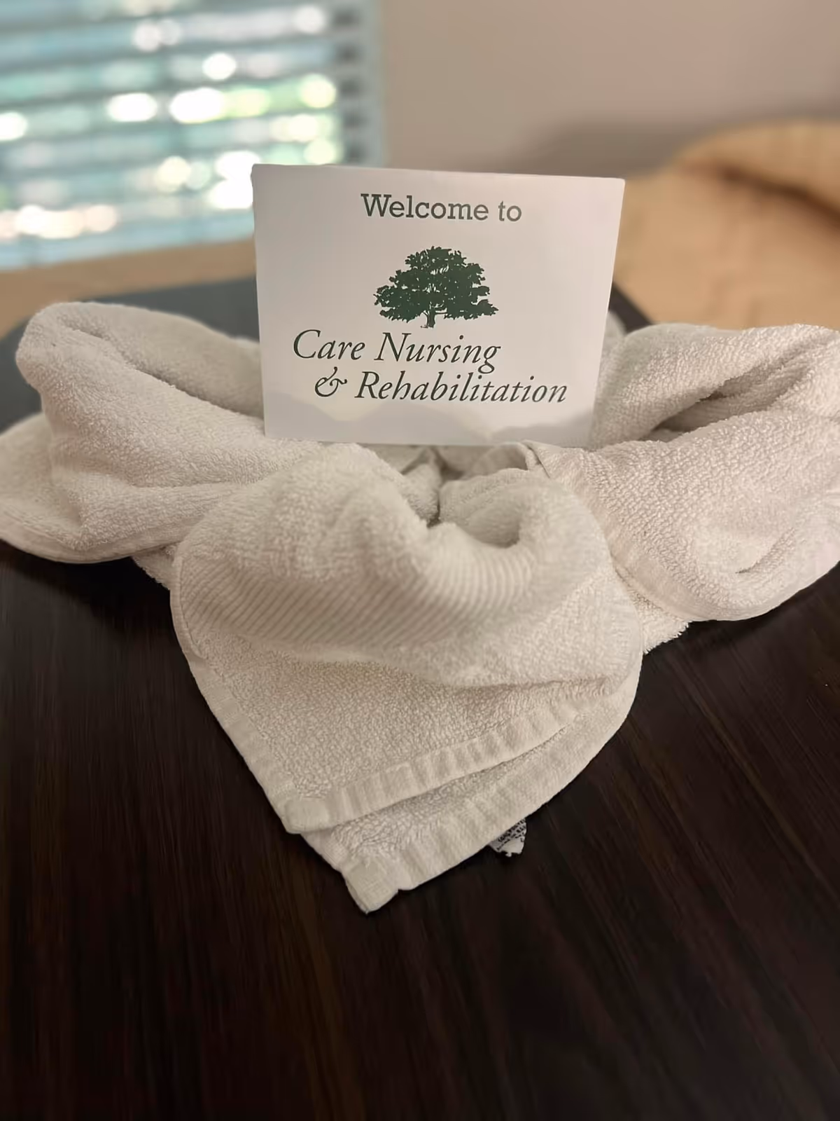 A white towel folded in a decorative shape on a dark wooden surface with a card placed in the center that reads 'Welcome to Care Nursing & Rehabilitation' with a tree logo above the text.
