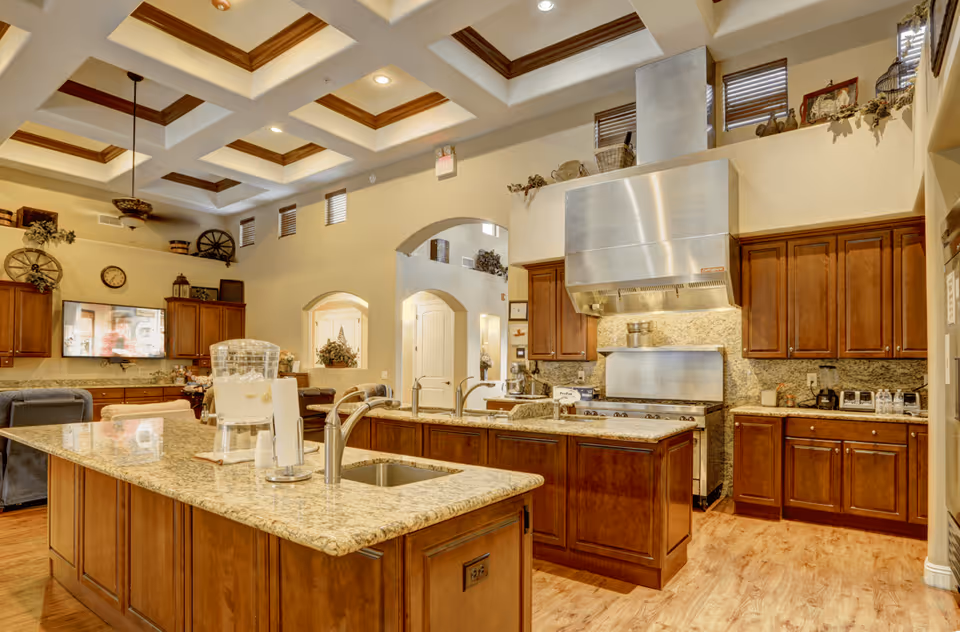 Spacious kitchen area with wooden cabinets, granite countertops, and a large island with a sink and water dispenser. The kitchen features a stainless steel range hood and stove, with decorative items on top of the cabinets. The ceiling has a coffered design with recessed lighting, and there is a living area visible in the background with a wall-mounted TV and comfortable seating.