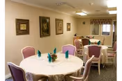 Interior view of a dining room with round tables covered with white tablecloths, each set with teal napkins and small floral centerpieces. The room has beige walls adorned with framed artwork and carpeted flooring. Several pink cushioned chairs are arranged around the tables.