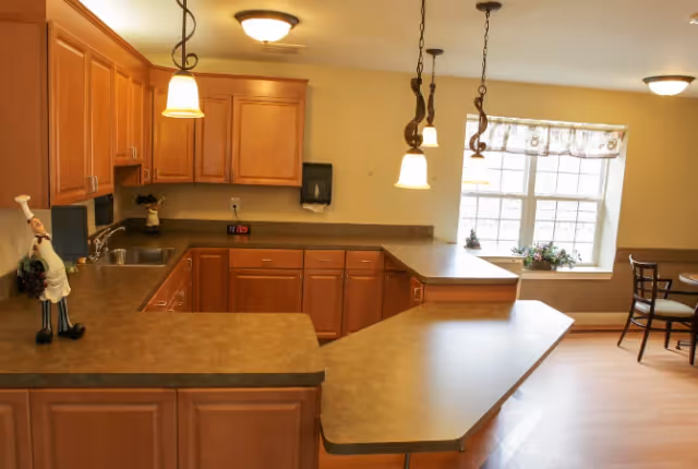 A kitchen area with wooden cabinets, a countertop with a unique angled extension, three hanging pendant lights, a window with a valance, and a small dining table with chairs near the window. There is a decorative chef figurine on the counter and a clock on the back counter showing 7:09.