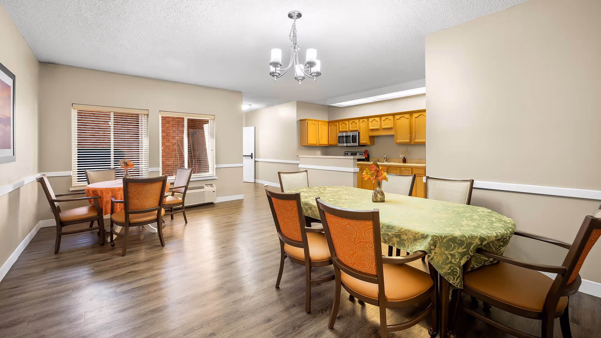 A spacious dining area with two tables covered with tablecloths, surrounded by chairs. The room has wooden flooring, beige walls, and a chandelier hanging from the ceiling. In the background, there is a kitchen area with wooden cabinets, a microwave, and a refrigerator. Two windows with blinds let in natural light.