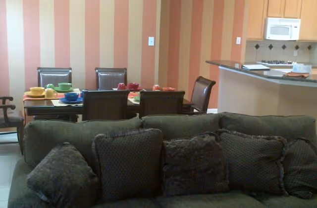 Interior view of a living and dining area in a senior living facility. In the foreground, there is a green sofa with multiple cushions. Behind the sofa, there is a dining table set with colorful bowls and cups, surrounded by six chairs. The wall behind the dining table has vertical orange and beige stripes. To the right, a kitchen counter with a granite countertop and a microwave mounted above a stove is visible.