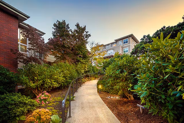 A paved walkway with a metal handrail curves through a landscaped garden area with dense green shrubs and trees. On the left, there is a red brick building with windows, and in the background, a multi-story residential building is visible under a clear sky during early evening.