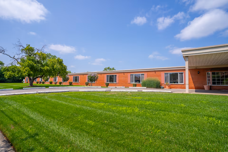Exterior view of a single-story brick building with several windows, a covered entrance on the right, and a well-maintained green lawn with a tree and shrubs in front under a partly cloudy blue sky.