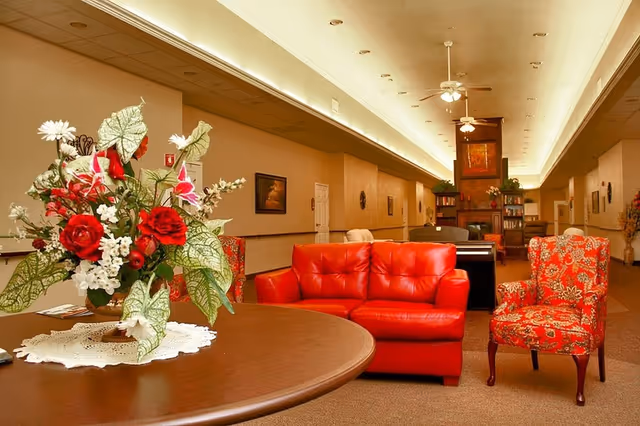 Interior view of a senior living facility lounge area with a round wooden table holding a floral arrangement with red and white flowers in the foreground. Behind the table is a red leather sofa and a red patterned armchair. The room has beige walls, ceiling fans, recessed lighting, and a fireplace with bookshelves in the background.