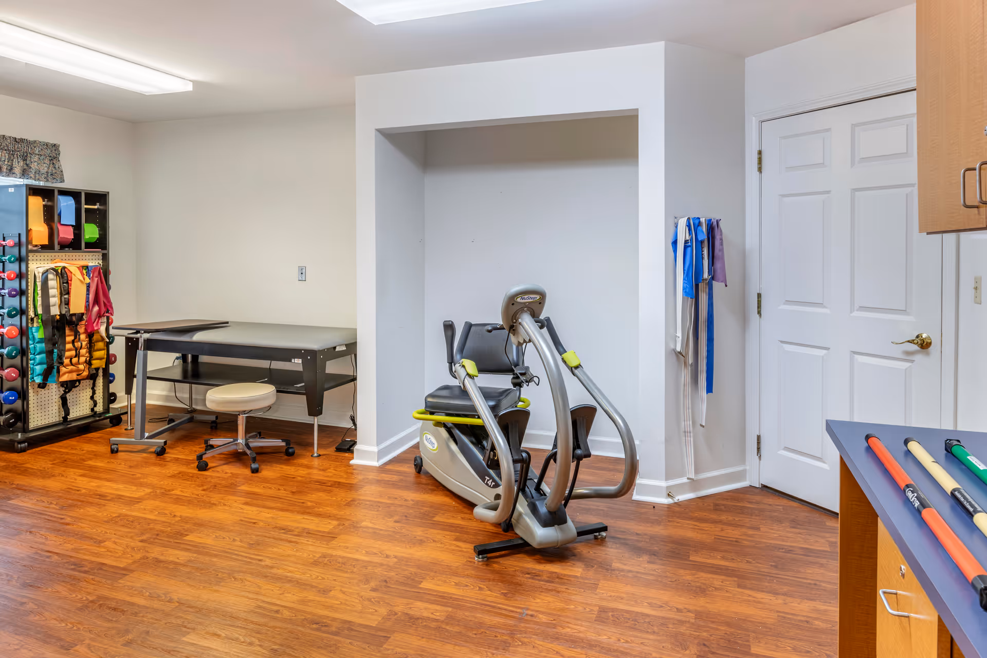 A room with wooden flooring containing exercise equipment including a recumbent exercise bike, a rack with colorful weights and resistance bands, a therapy table with a rolling stool, and a countertop with pool cues. The walls are white, and there is a closed white door on the right side.