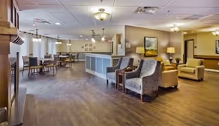 A spacious senior living facility common area with wooden flooring, several tables and chairs arranged for dining, and comfortable armchairs near a reception or service counter. The room is well-lit with ceiling lights and pendant lamps, and decorated with framed artwork and lamps on side tables.