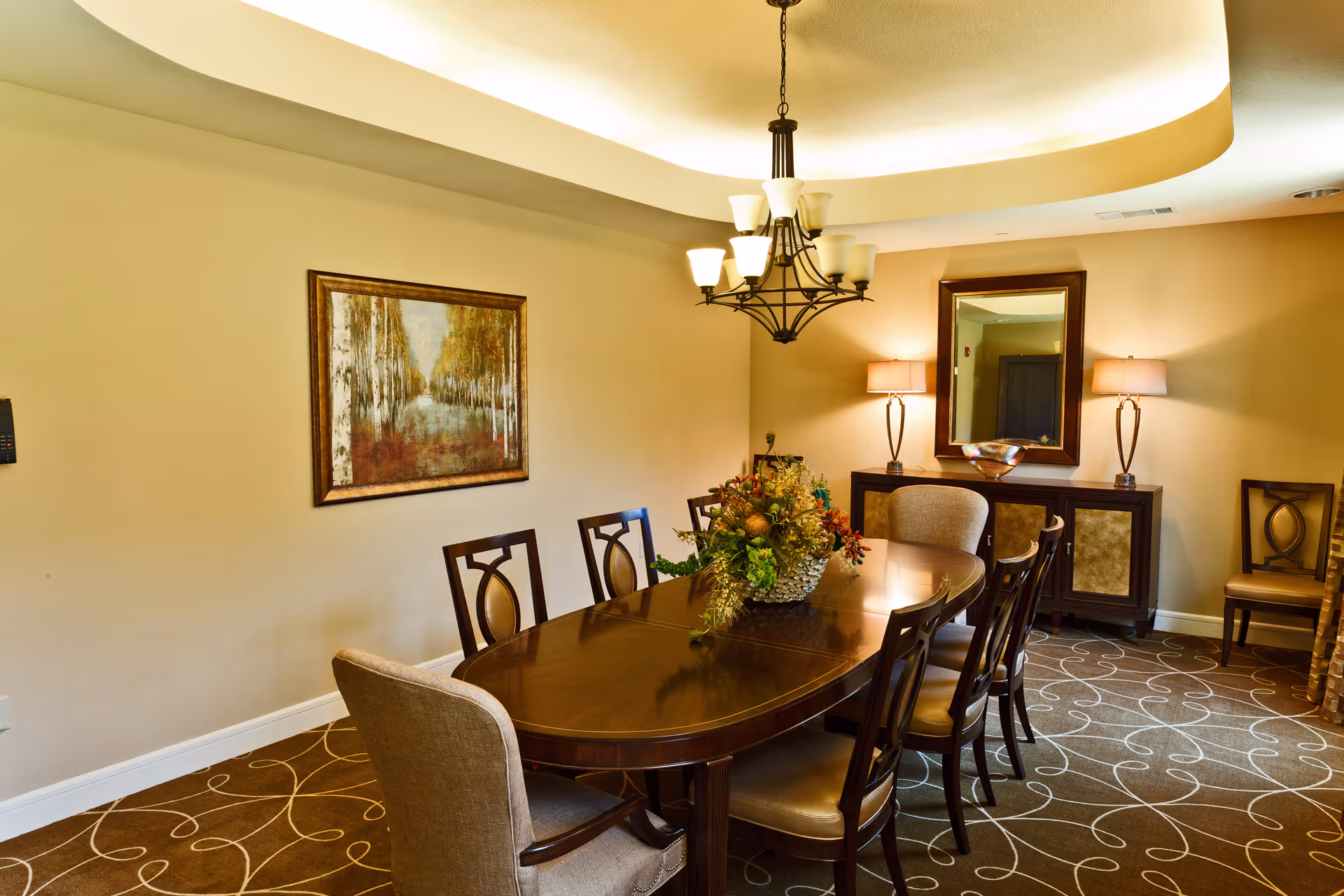 A well-lit dining room with a large oval wooden table surrounded by eight chairs, a floral centerpiece on the table, a framed painting on the wall, a sideboard with two lamps and a mirror above it, and a patterned carpet on the floor.